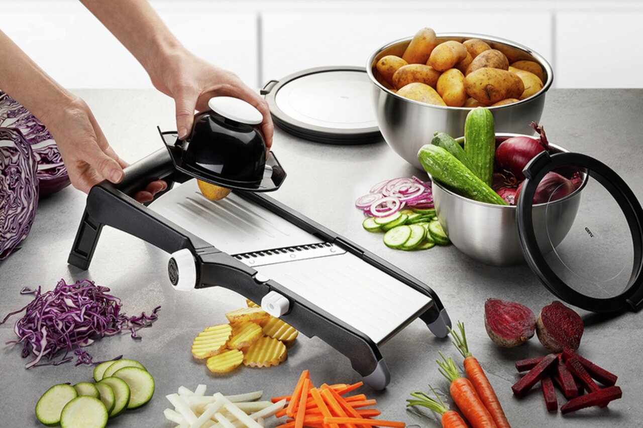Gemüsehobel Laola in seitlicher Perspektive, umgeben von geschnittenem Gemüse wie Karotten, Gurken und Kartoffeln.