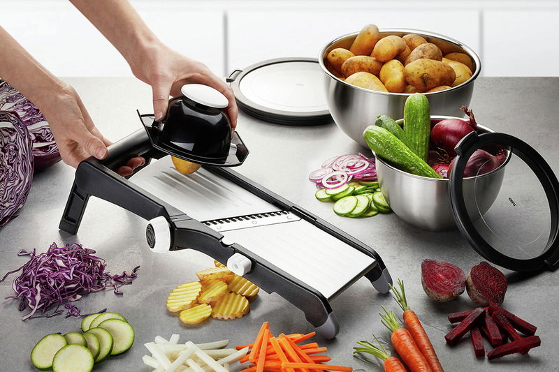 Gemüsehobel Laola in seitlicher Perspektive, umgeben von geschnittenem Gemüse wie Karotten, Gurken und Kartoffeln.