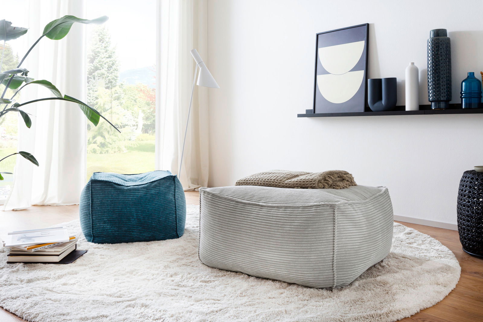 Zwei quadratische Hocker in einem modernen Wohnzimmer, einer in Grau und einer in Blau, auf einem runden Teppich vor einem großen Fenster. Perspektive von schräg oben.
