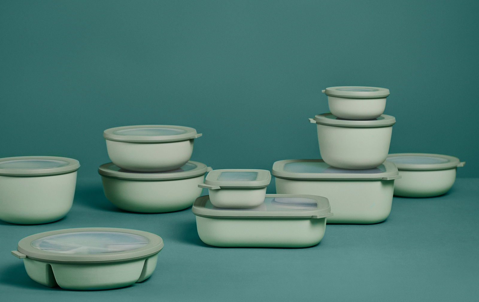 Mehrere grüne Bento-Schüsseln und -Behälter in verschiedenen Größen, gestapelt und nebeneinander auf einer grünen Oberfläche, aus seitlicher Perspektive fotografiert.