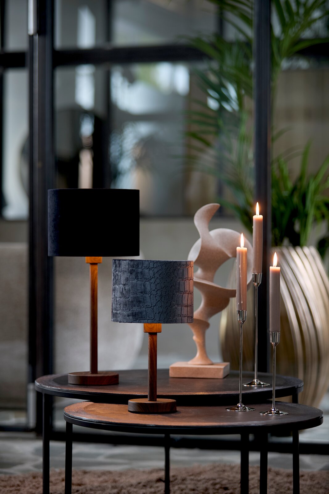 Twee stijlvolle tafellampen met een houten voet en verschillende lampenkappen, de ene in het zwart en de andere met een krokodillenleerpatroon, op ronde tafels in een modern interieur. Zijaanzicht.