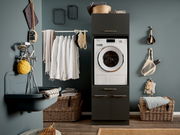 Hauswirtschaftsraum mit Waschmaschine in einem dunklen Schrank, umgeben von Körben und einem Waschbecken. Frontalansicht.