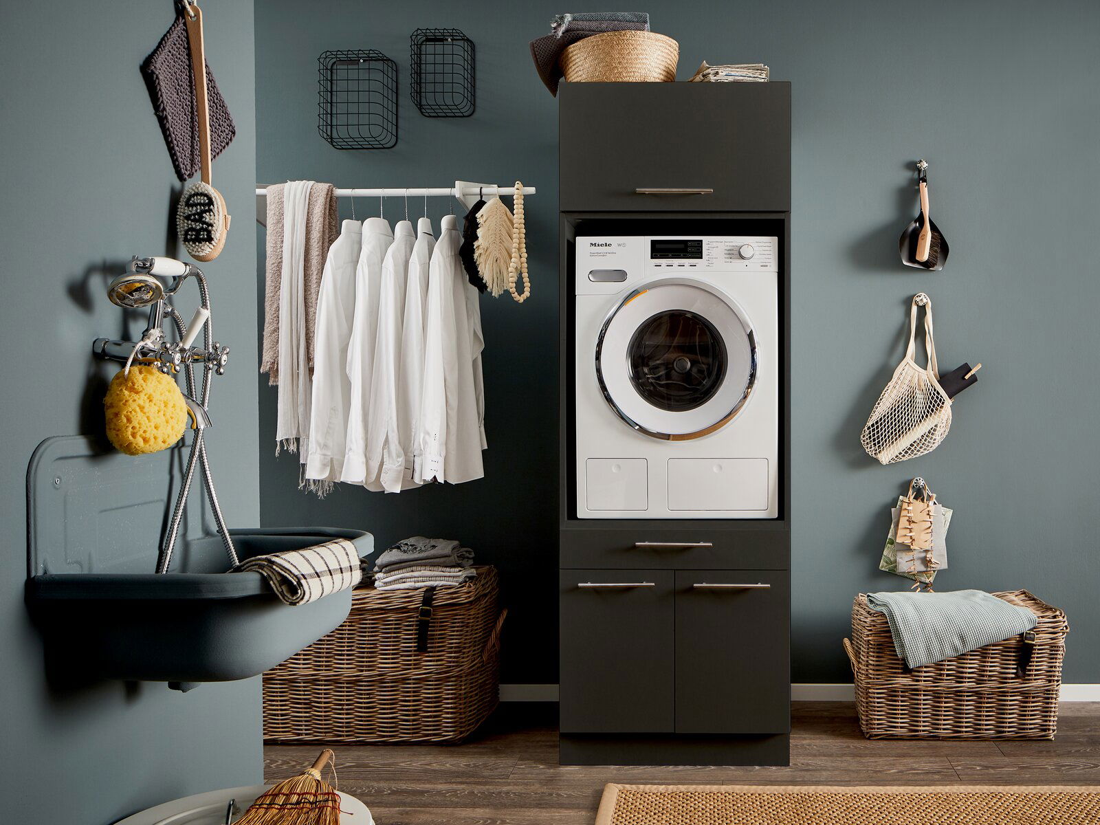 Hauswirtschaftsraum mit Waschmaschine in einem dunklen Schrank, umgeben von Körben und einem Waschbecken. Frontalansicht.