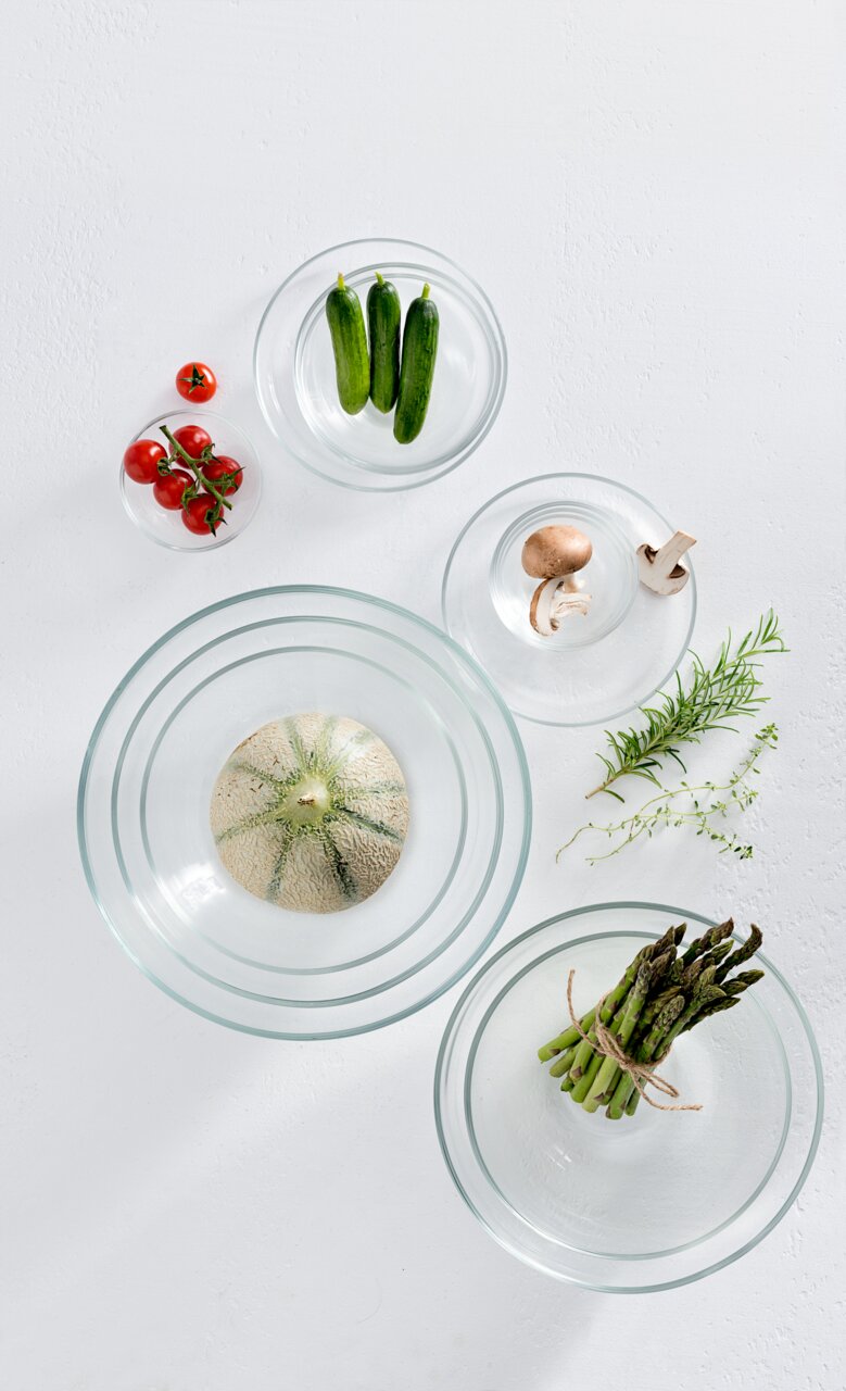 Vogelperspektive auf mehrere Glasschalen mit frischem Gemüse wie Tomaten, Gurken, Pilzen, Spargel und einer Melone.