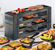 Perspektive von schräg vorne: Ein schwarzes Pizza-Raclette-Gerät mit mehreren Ebenen, auf denen Pfännchen mit verschiedenen Zutaten wie Gemüse und Käse zu sehen sind. Auf der oberen Grillfläche liegen Würstchen und Gemüse.