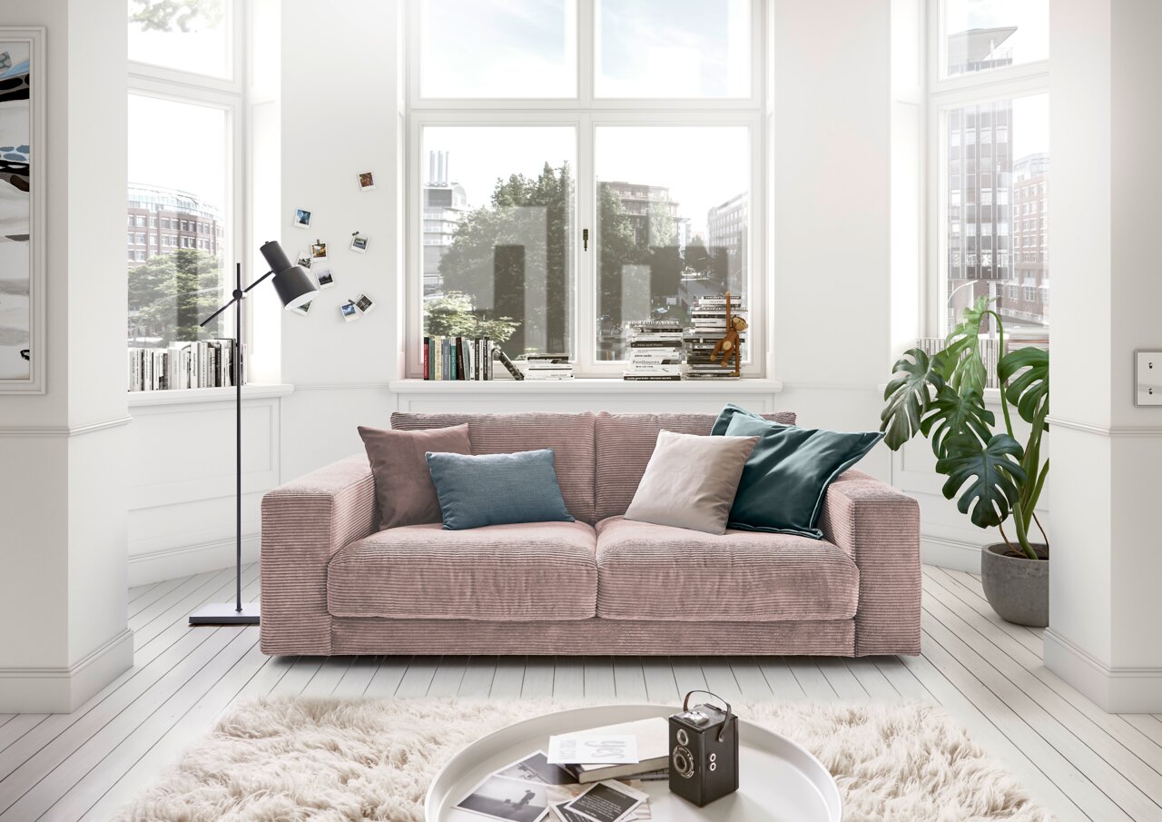 Lounge Handmade Sofa 2-Sitzer LH-SAMBIA Gemütliches 2-Sitzer-Sofa in einem hellen Wohnzimmer mit großen Fenstern, dekoriert mit bunten Kissen, aus der Frontalperspektive.