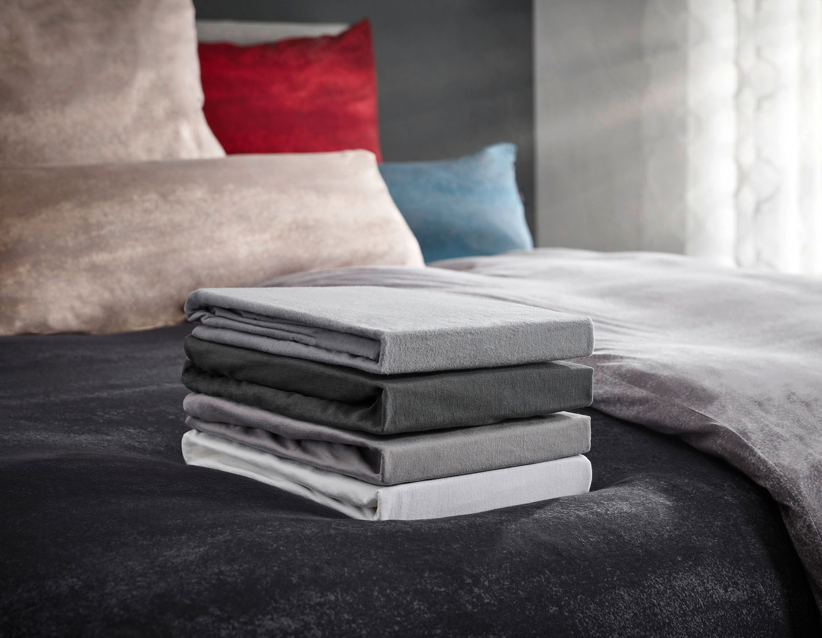 Novel Topper-Spannbetttuch Florenz 180x200cm Ein Stapel von vier gefalteten Spannbetttüchern in verschiedenen Grautönen auf einem Bett, fotografiert aus einer seitlichen Perspektive.