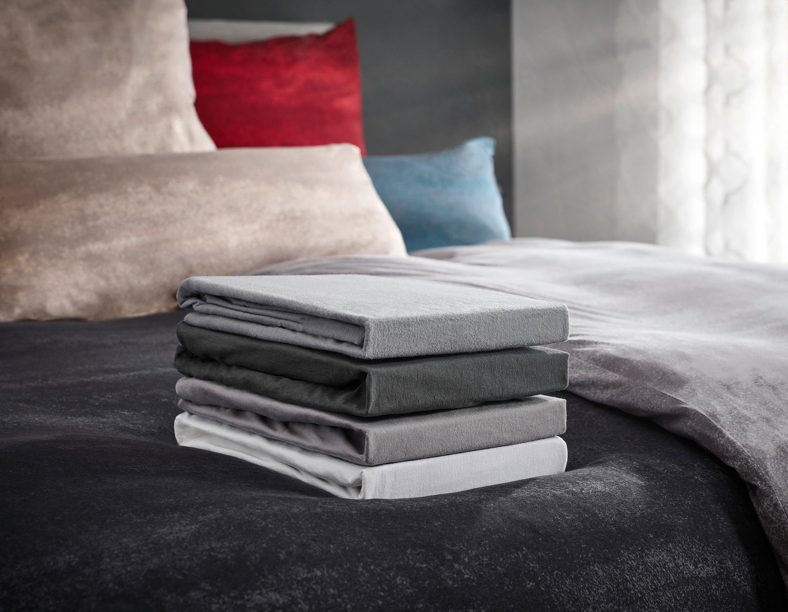 Novel Topper-Spannbetttuch Florenz 180x200cm Ein Stapel von vier gefalteten Spannbetttüchern in verschiedenen Grautönen auf einem Bett, fotografiert aus einer seitlichen Perspektive.