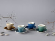 Vier farbige Espressotassen mit passenden Untertassen aus Keramik (Beige, Hellblau, Dunkelblau, Mintgrün) auf grauem Hintergrund mit dekorativen Trockenpflanzen und Kieselsteinen, frontal aus leicht erhöhter Perspektive