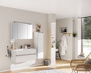 Moderner Badezimmer-Mittelschrank in Weiß, rechts an der Wand montiert, mit minimalistischer Einrichtung und natürlichem Lichteinfall von der Seite.