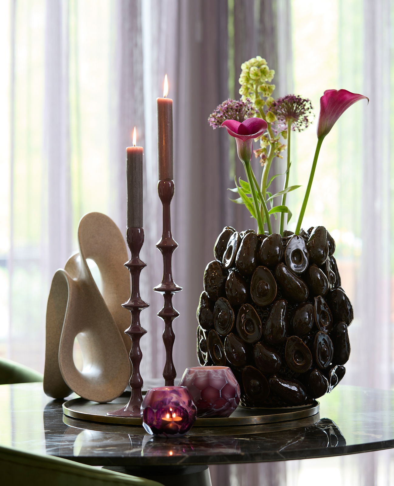 Dekorative Tischszene mit einer antiken Schale, die als Unterlage für moderne Kerzenhalter und eine skulpturale Vase mit Blumen dient. Die Perspektive zeigt die Objekte von der Seite auf einem glänzenden Tisch.