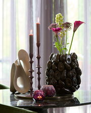 Dekorative Tischszene mit einer antiken Schale, die als Unterlage für moderne Kerzenhalter und eine skulpturale Vase mit Blumen dient. Die Perspektive zeigt die Objekte von der Seite auf einem glänzenden Tisch.