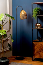 Staande zwarte booglamp met warm verlichte, gevlochten metalen kap, vanuit vooraanzicht gefotografeerd in een stijlvol interieur met planten en een houten kast.