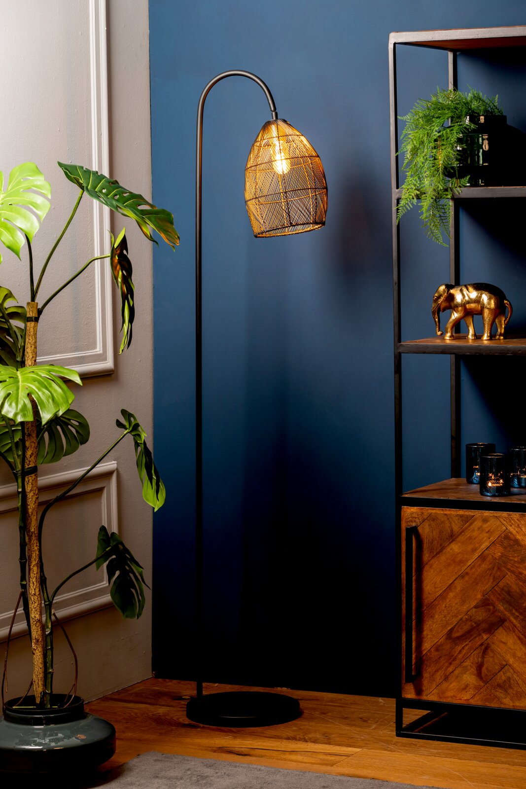 Staande zwarte booglamp met warm verlichte, gevlochten metalen kap, vanuit vooraanzicht gefotografeerd in een stijlvol interieur met planten en een houten kast.