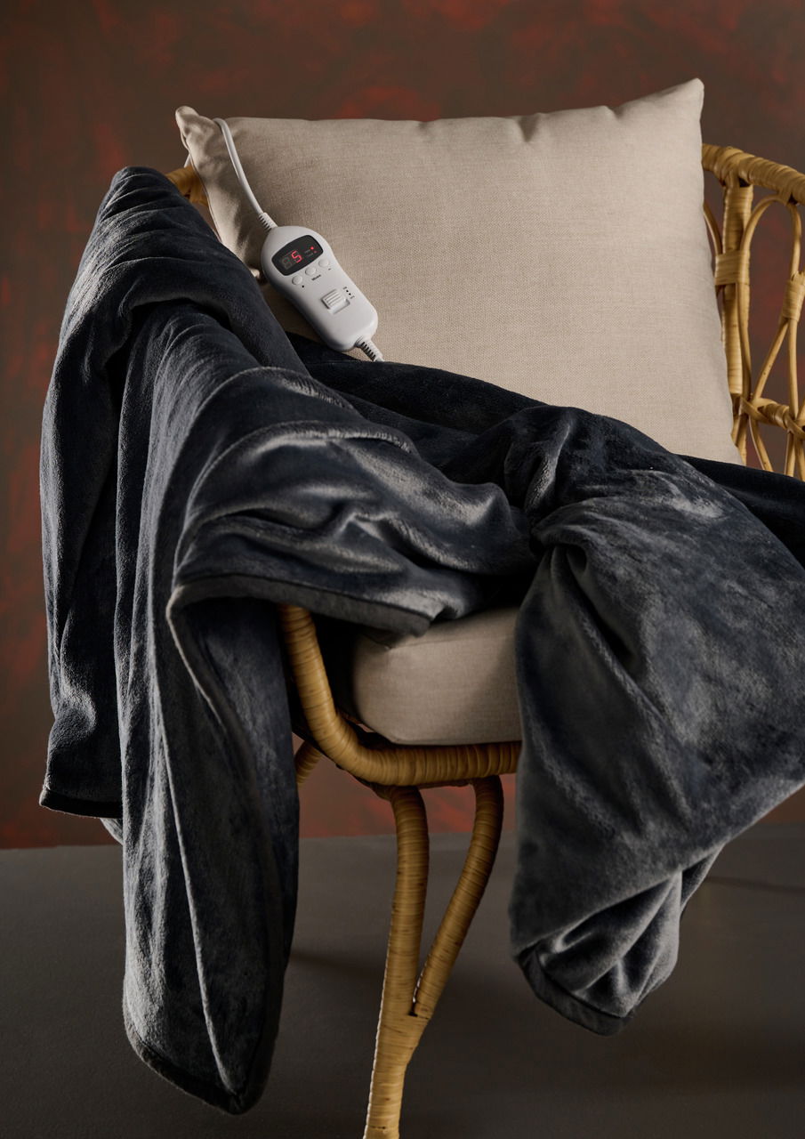 Day elektr. Wärmedecke Elektrische Wärmedecke in Dunkelgrau auf einem Stuhl drapiert, mit Fernbedienung auf der Sitzfläche, seitliche Perspektive.