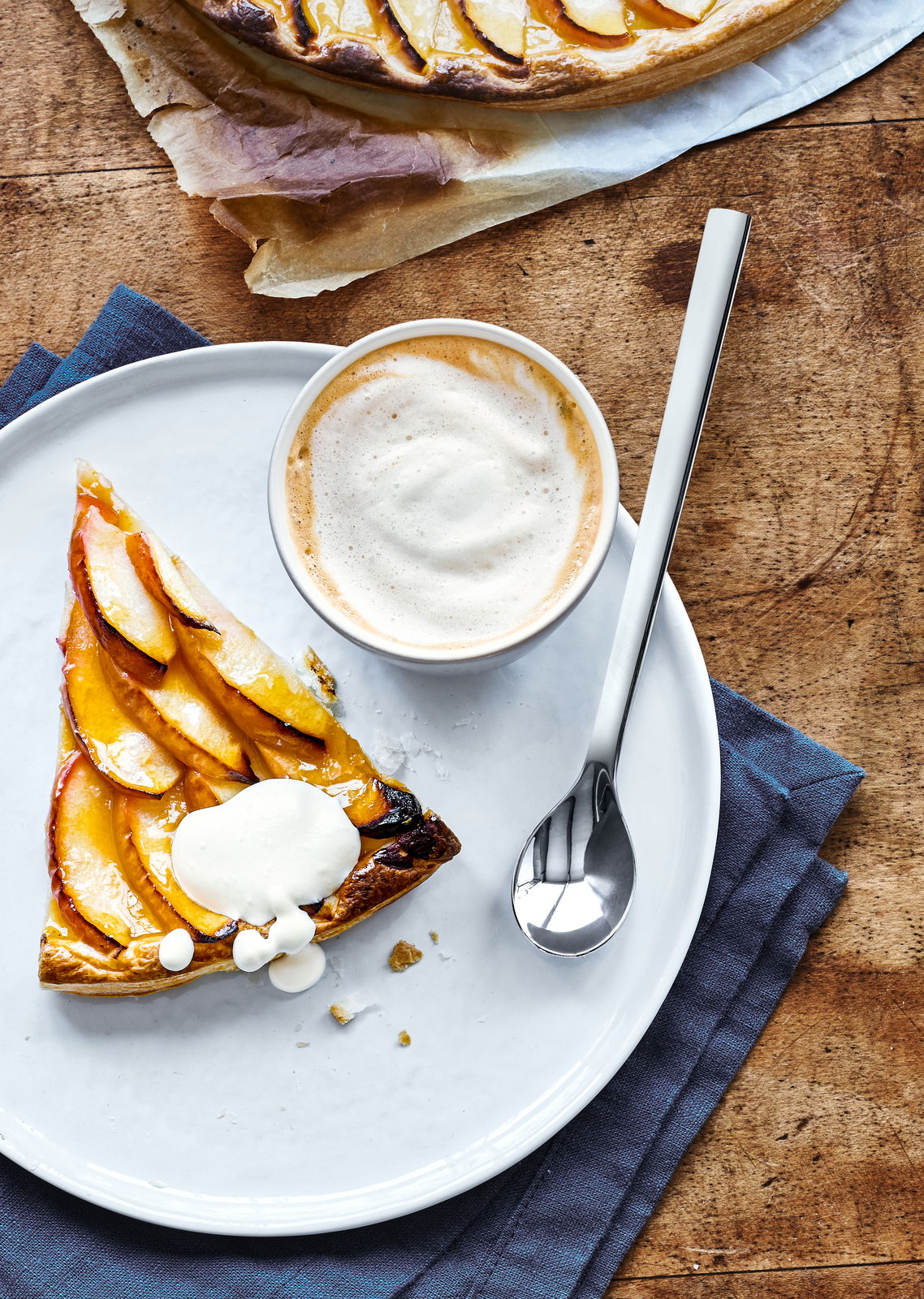 WMF Kaffeebecherlöffel 6-tlg. NUOVA Kaffeebecherlöffel aus Edelstahl neben einer Tasse Cappuccino und einem Stück Obstkuchen auf einem weißen Teller, von oben betrachtet.