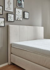 Beiges Boxspringbett mit gepolstertem Kopfteil, seitliche Perspektive, dekoriert mit gerahmten Bildern an der Wand.