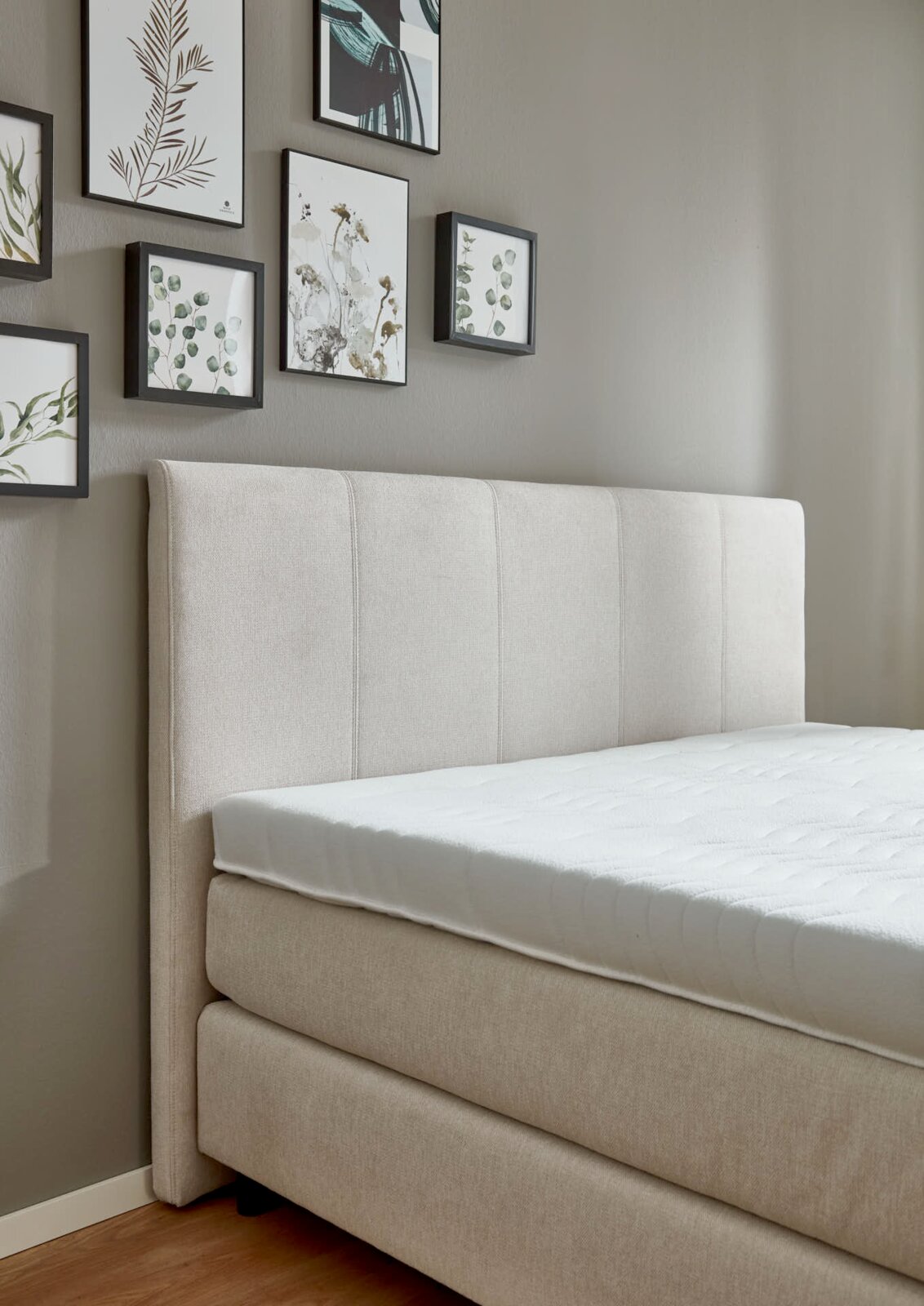 Beige boxspringbed met gestoffeerd hoofdeinde, gezien vanaf de zijkant, met ingelijste foto’s aan de muur.