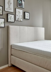 Beige boxspringbed met gestoffeerd hoofdeinde, gezien vanaf de zijkant, met ingelijste foto’s aan de muur.