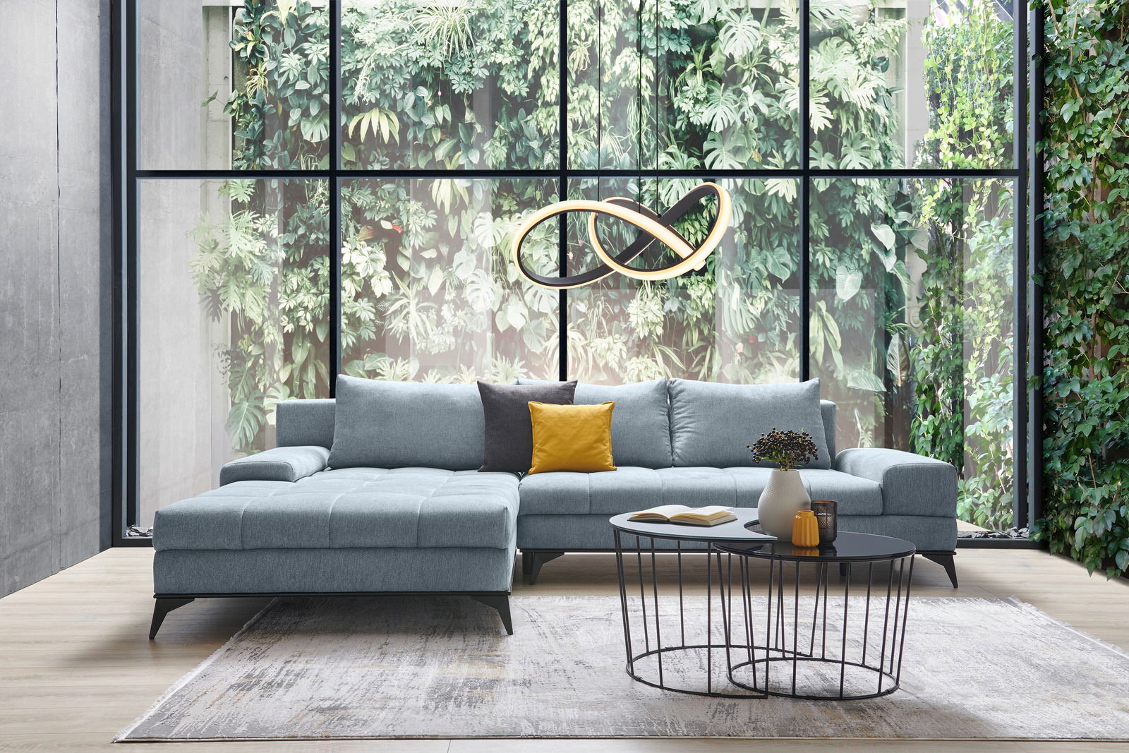 Moderne Rundecke Ottli in hellgrauem Stoff, mit gelbem und grauem Kissen, vor einer großen Fensterfront mit Blick auf eine grüne Pflanzenwand, aus der Frontalperspektive fotografiert.