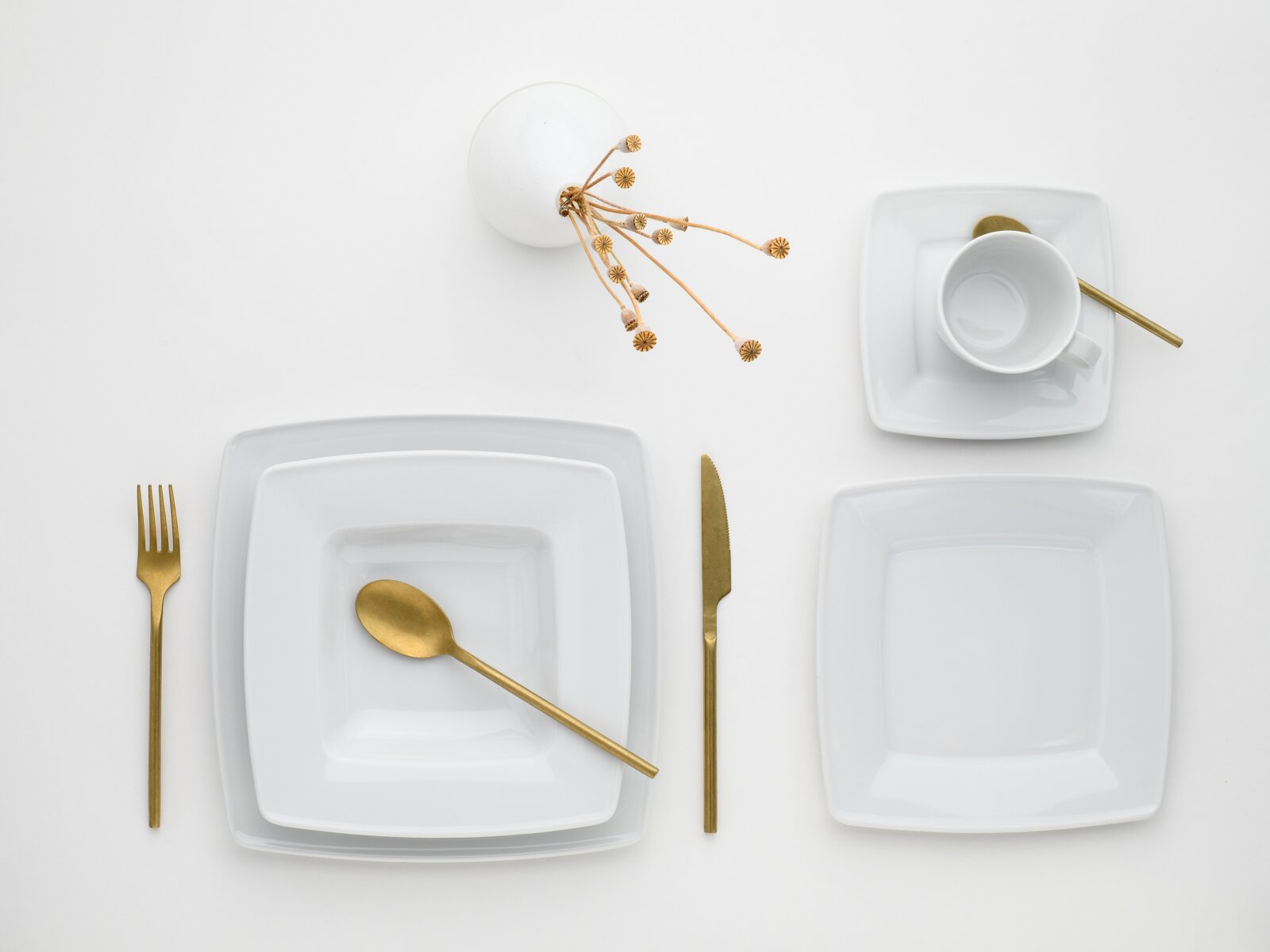 Minimalistisches Tischgedeck in Draufsicht (Top-Down): weiße quadratische Teller und Tasse mit Untertasse, goldfarbenes Besteck (Gabel, Messer, Löffel) und kleine Dekozweige auf weißem Hintergrund