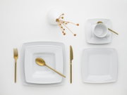 Minimalistisches Tischgedeck in Draufsicht (Top-Down): weiße quadratische Teller und Tasse mit Untertasse, goldfarbenes Besteck (Gabel, Messer, Löffel) und kleine Dekozweige auf weißem Hintergrund