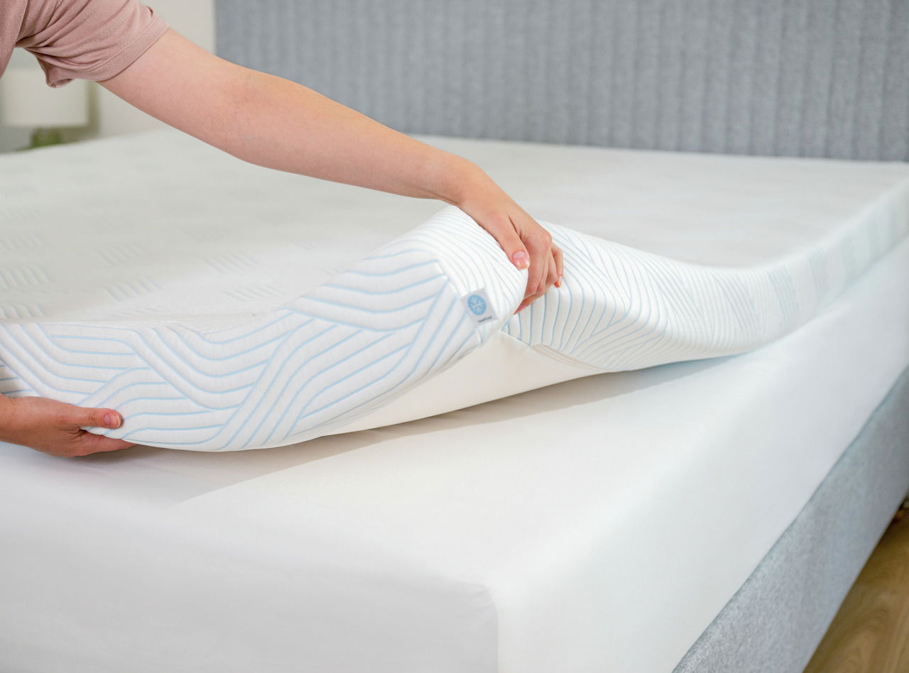 Nahaufnahme eines Toppers, der von einer Hand angehoben wird, um die weiche und flexible Struktur zu zeigen. Der Topper hat ein gestreiftes Muster und liegt auf einer Matratze.