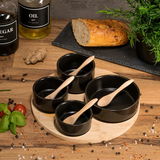 Servierset aus Bambus mit vier schwarzen Schalen und Holzlöffeln, von oben fotografiert, umgeben von Gewürzen, Brot und Pflanzen.