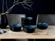 Schwarze Keramikschalen mit blau glasierter Innenseite, teils gestapelt und mit Löffel, auf Holzbrett auf einem Holztisch vor dunklem Hintergrund, Perspektive: frontale leicht erhöhte Ansicht