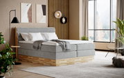 Een modern boxspringbed in een stijlvolle slaapkamer, gezien vanaf de zijkant, met een grijze stoffen bekleding en houten details, omgeven door een elegante inrichting en grote ramen.