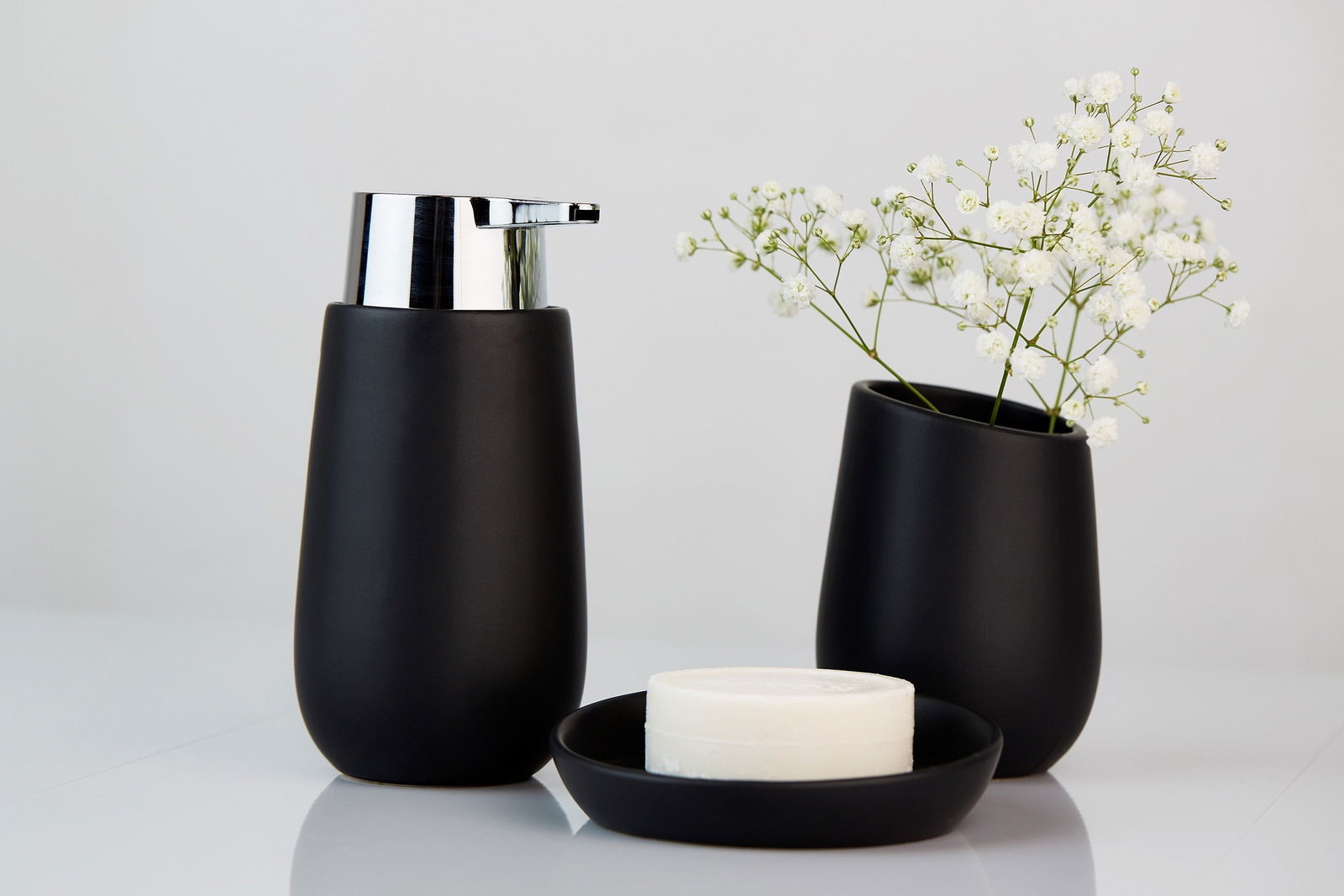 Schwarzer Keramik-Seifenspender mit glänzendem Chrom-Pumpkopf, frontal fotografiert, neben einer passenden Vase mit weißen Blumen und einer Seifenschale.