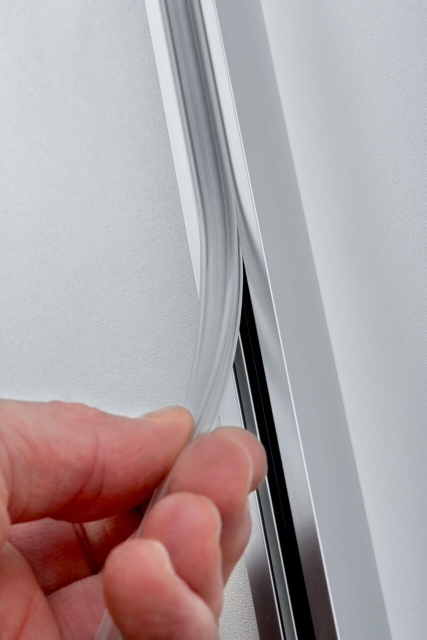PAULMANN SavetyCoverStrip URAIL Nahaufnahme einer Hand, die eine transparente Sicherheitsabdeckung in eine Metallschiene einfügt, seitliche Perspektive.