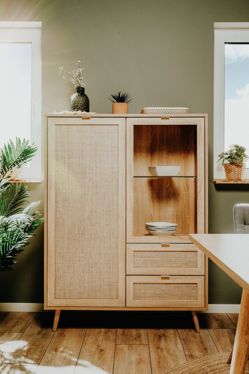 Hochwertiges Highboard aus Holz mit Rattan-Elementen, dekoriert mit Pflanzen und Geschirr, in einem modernen Raum mit Tageslicht von der Seite.