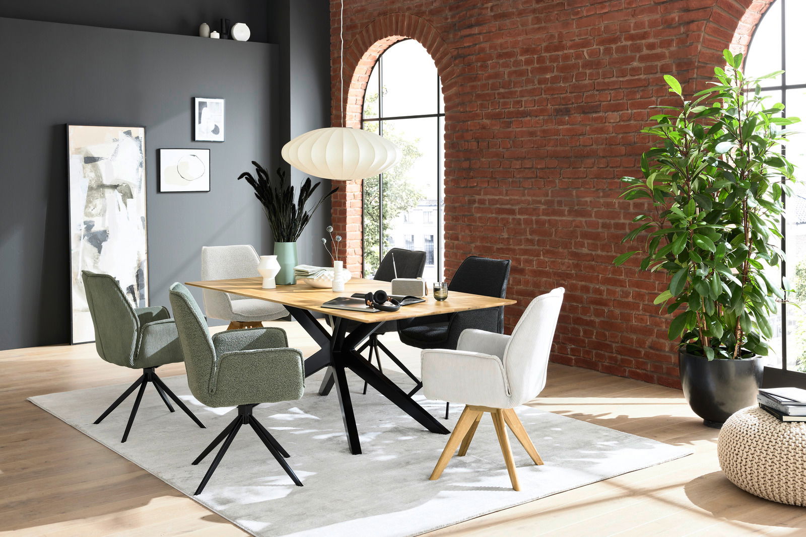 Moderner Essbereich mit einem Holztisch und verschiedenen Armlehnstühlen in einem stilvollen Raum mit Backsteinwand und großen Fenstern, seitliche Perspektive.