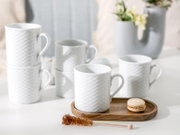Weiße Keramik-Kaffeetassen mit feiner Reliefstruktur und Henkel auf hellem Tisch, eine Tasse auf Holztablett mit Macaron; Perspektive: leicht erhöhte Frontansicht.