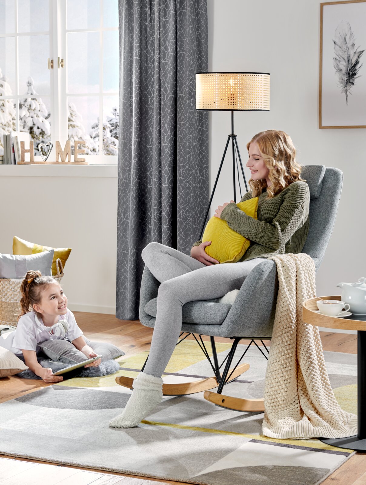 Een gezellige schommelstoel in een moderne woonkamer, gezien vanuit de zijkant, met een vrouw die een kussen vasthoudt en een kind dat op de grond zit.