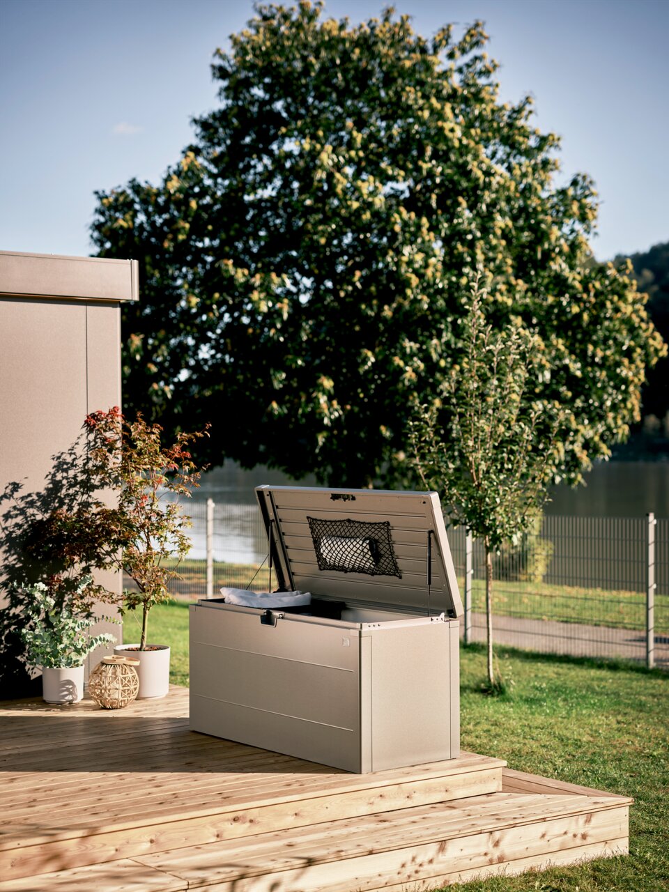 Biohort Stylebox 140 Beige metalen opbergbox voor buiten met open deksel en gaasvak, op een houten terras in de tuin, perspectief: licht zijdelings vooraanzicht
