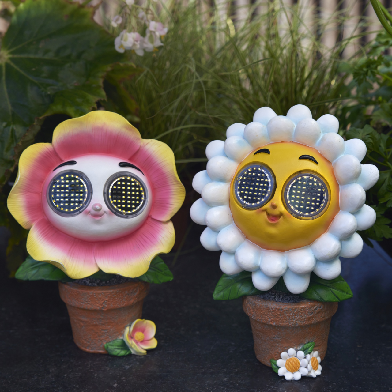 Zwei bunte Solarfiguren in Blumentöpfen, eine mit rosa Blütenblättern und eine mit weißen Blütenblättern, beide mit großen, runden Solaraugen, frontal fotografiert.