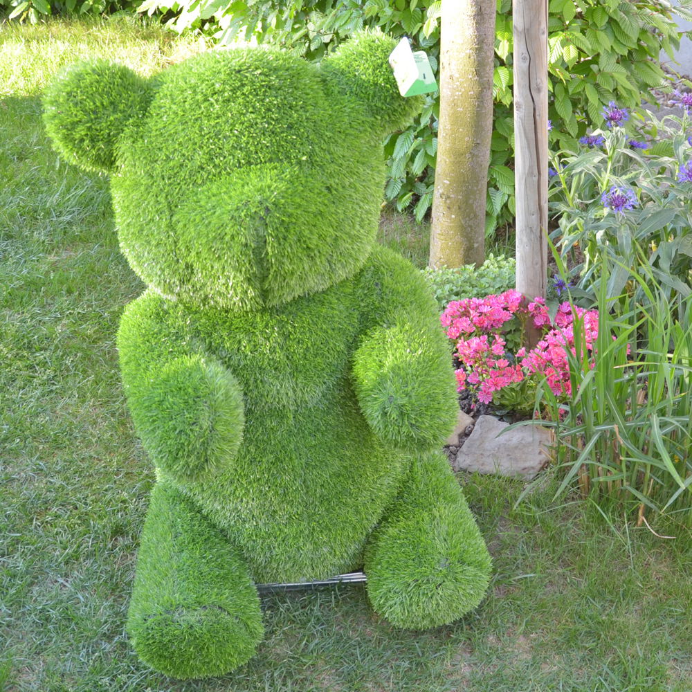 Grüner Bär aus Gras, frontal im Garten mit Blumen im Hintergrund