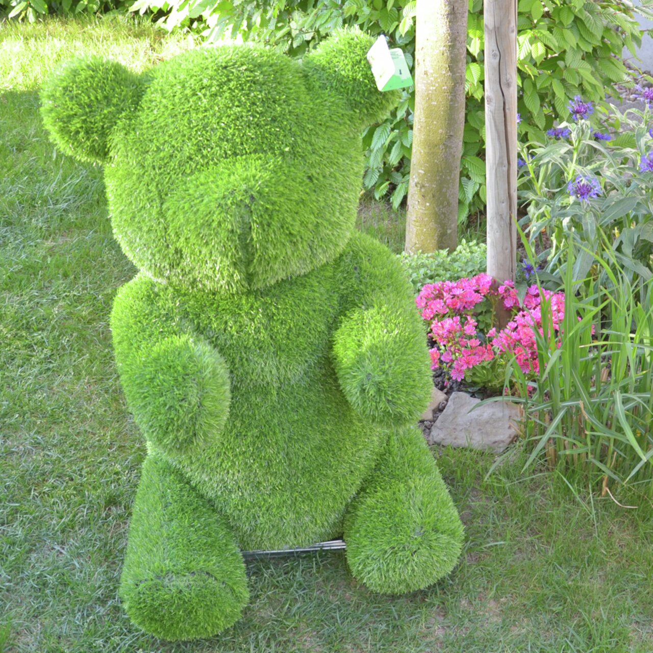 Grüner Bär aus Gras, frontal im Garten mit Blumen im Hintergrund