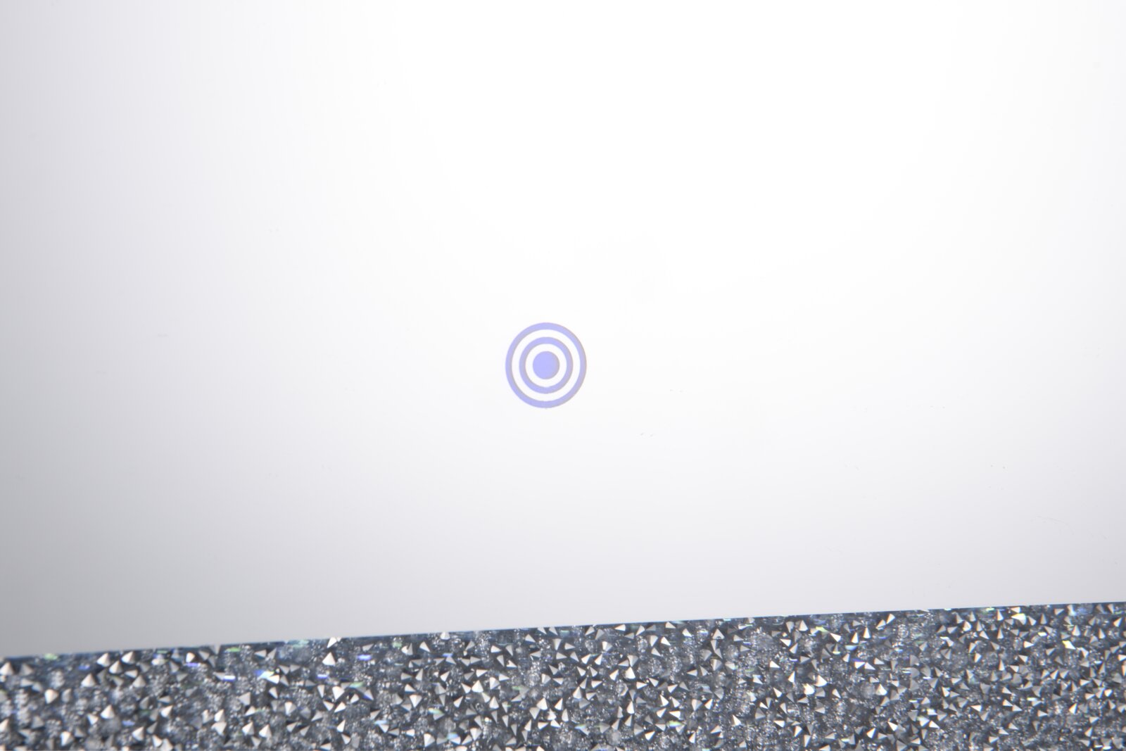 Weiße, minimalistische Produktoberfläche mit kleinem kreisförmigem Symbol in der Mitte und glitzerndem Rand am unteren Bildrand, Nahaufnahme in frontaler Draufsicht