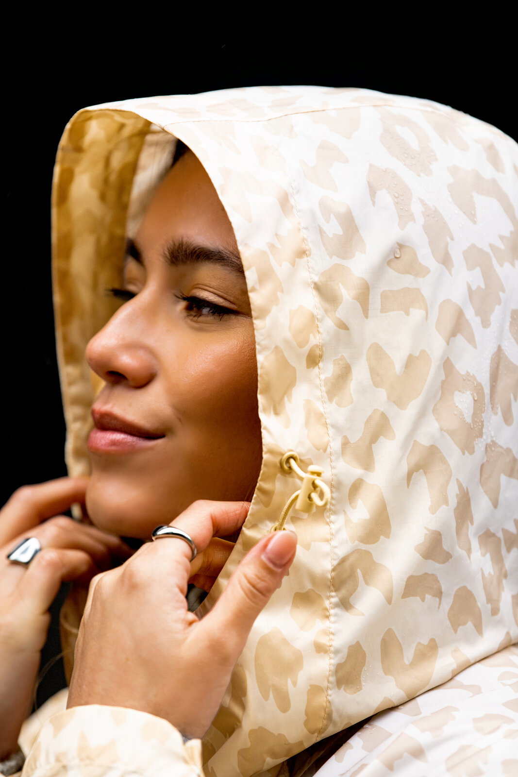 Nahaufnahme einer Frau im Profil, die die Kapuze einer beigen, gemusterten Regenjacke mit Kordelzug anpasst; Seitenansicht/Close-up.