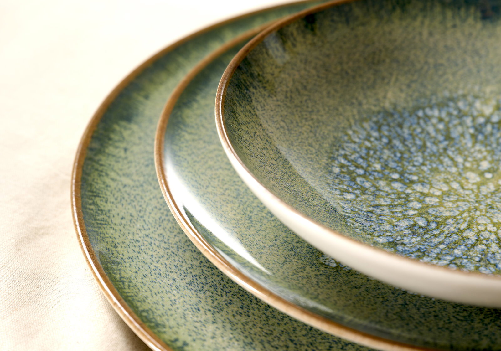 Nahaufnahme aus schräger Draufsicht auf zwei übereinanderliegende Keramik-Teller in Grün-Blau mit gesprenkelter Glasur und braunem Rand auf hellem Stoff