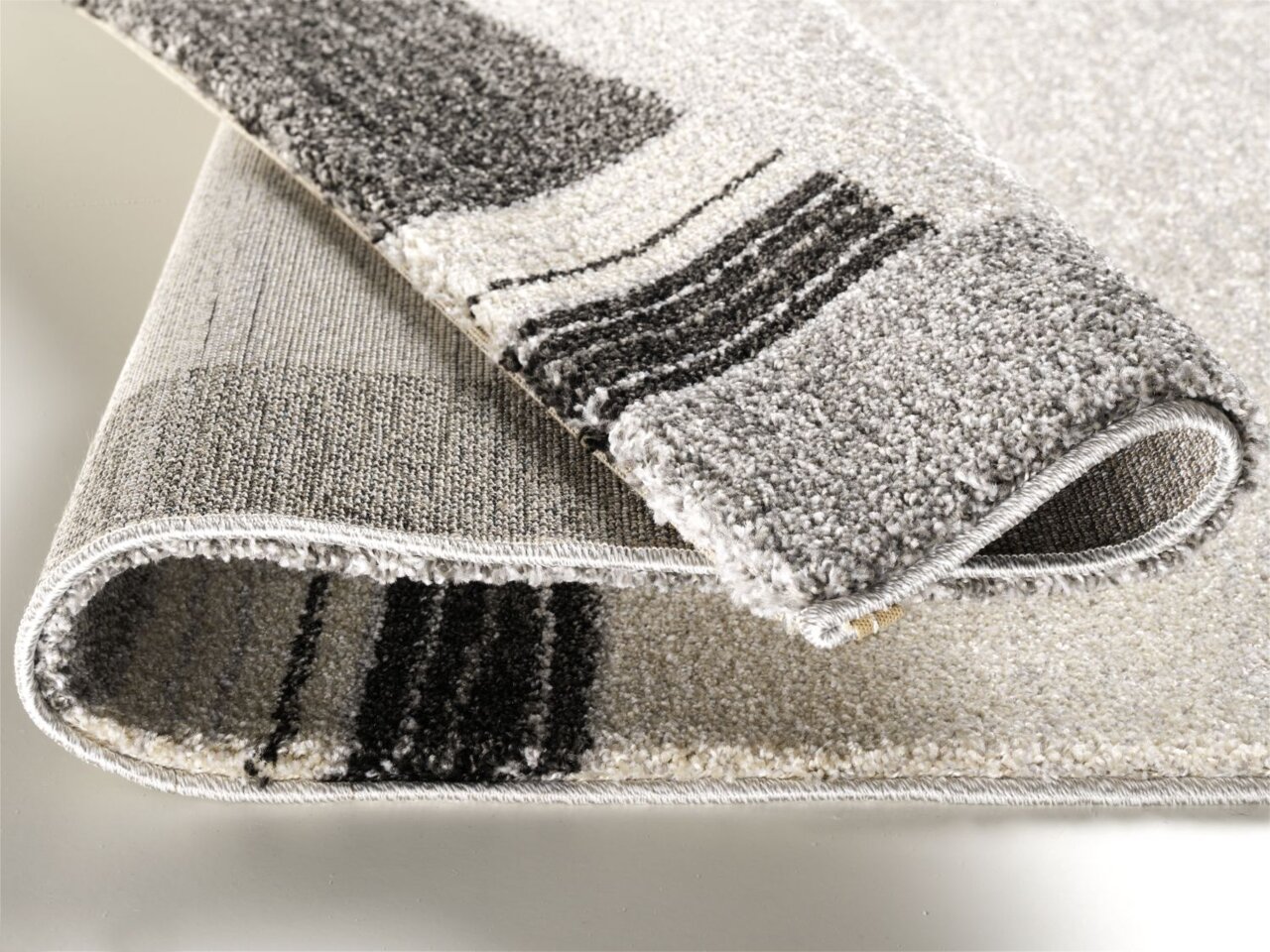 Nahaufnahme eines gefalteten Teppichs mit grauen und schwarzen Streifen, Perspektive von der Seite.