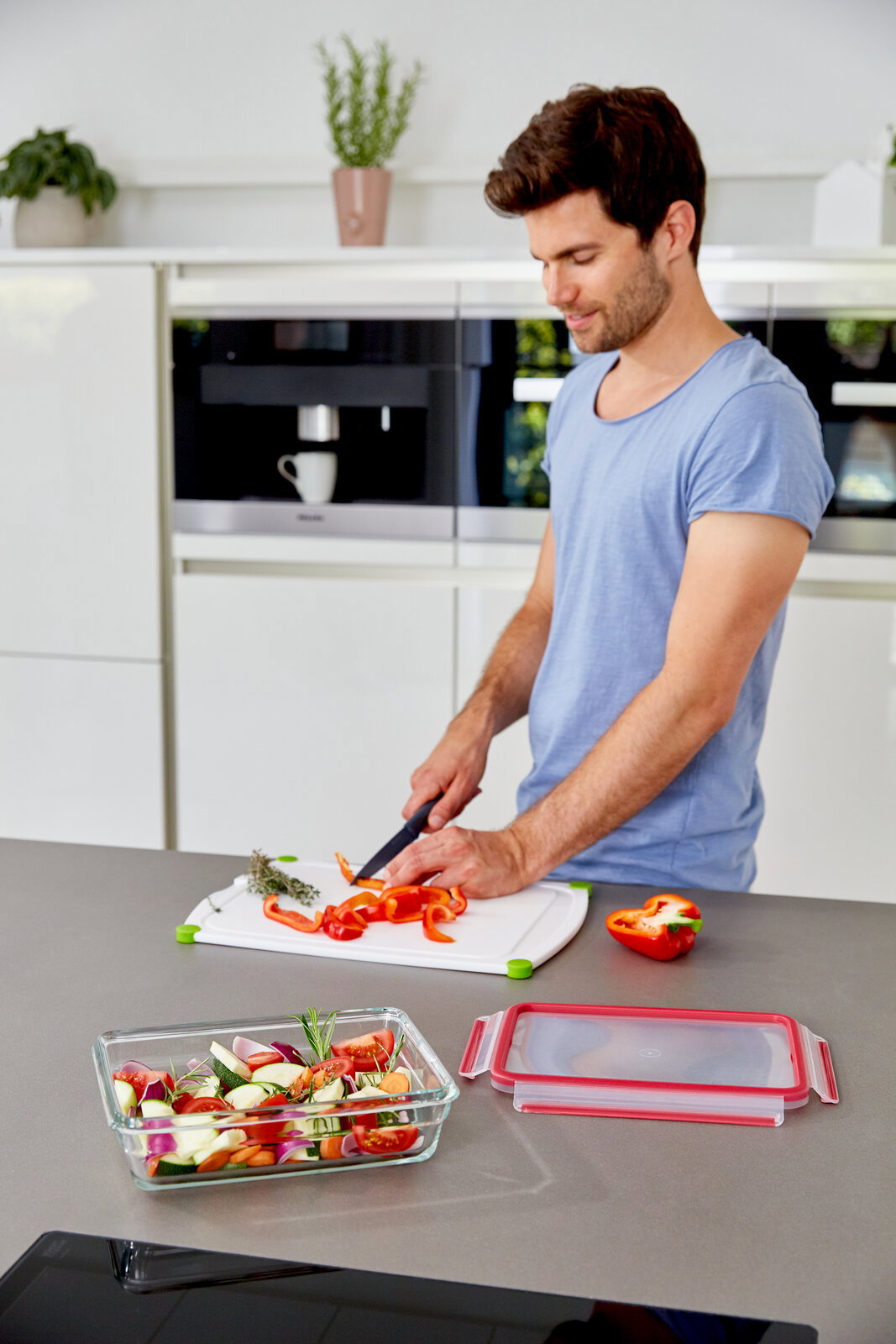 Mann schneidet Paprika in moderner Küche, im Vordergrund ein rechteckiges Glasbehälter mit frischem Gemüse, Perspektive von oben.
