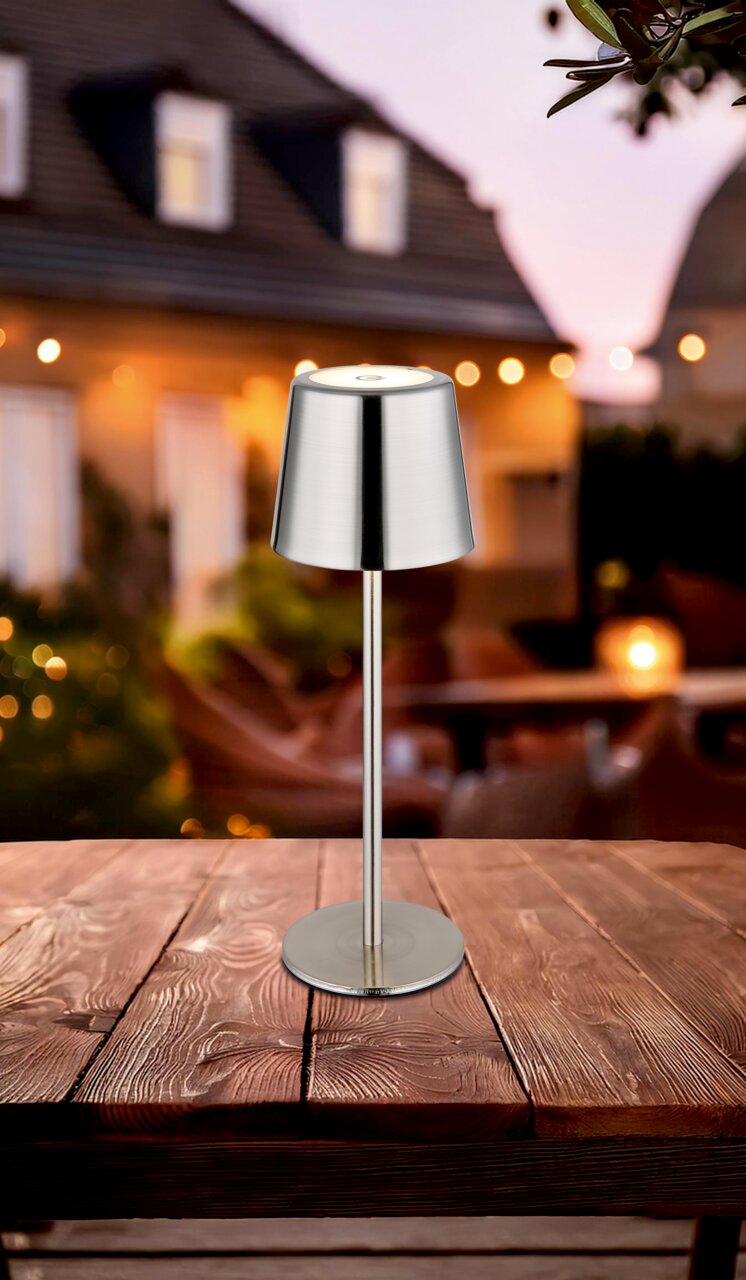 Moderne Tischleuchte aus Metall auf einem Holztisch, im Freien bei Dämmerung fotografiert.