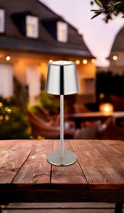 Moderne Tischleuchte aus Metall auf einem Holztisch, im Freien bei Dämmerung fotografiert.