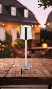 Moderne Tischleuchte aus Metall auf einem Holztisch, im Freien bei Dämmerung fotografiert.
