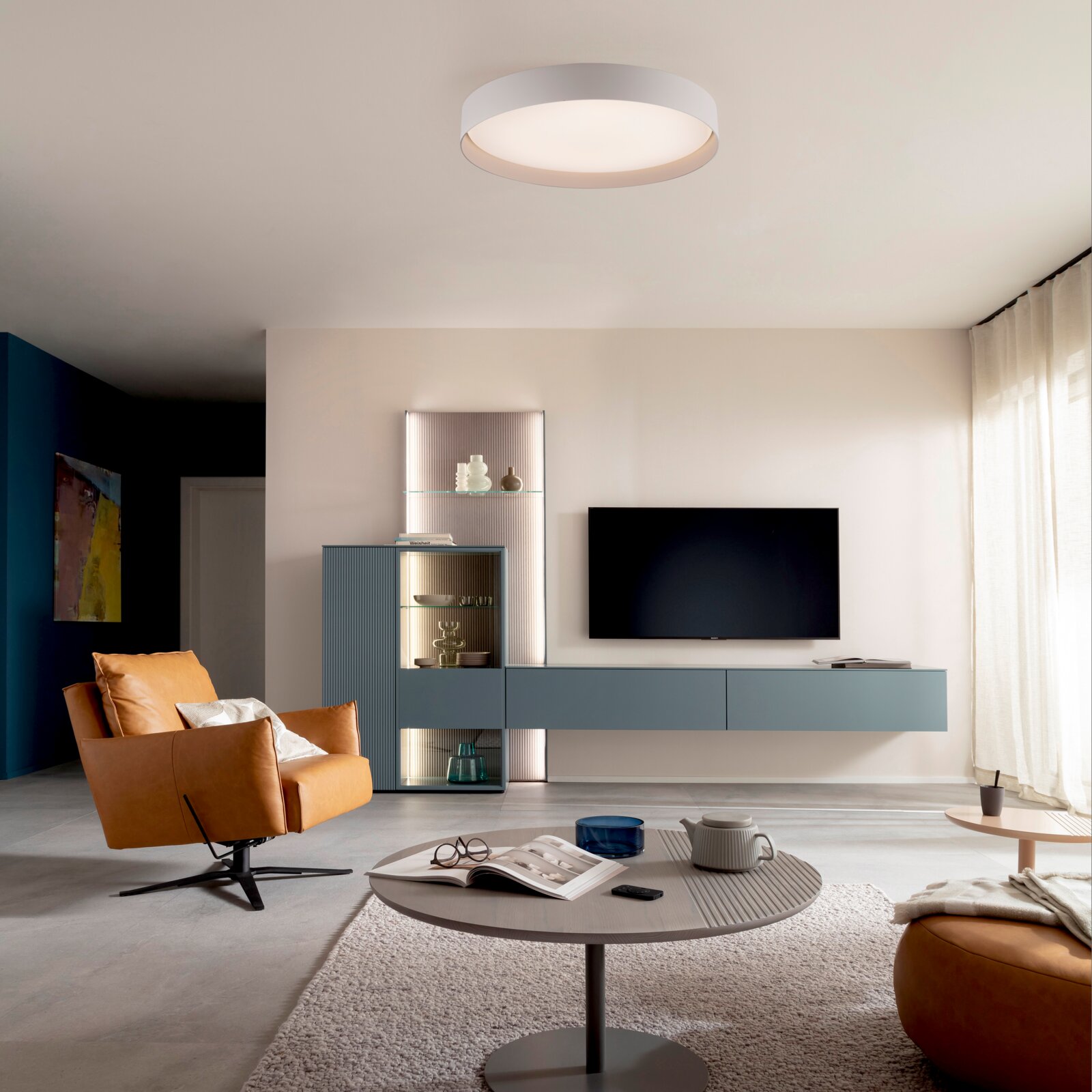 Modernes Wohnzimmer mit runder, weißer LED-Deckenleuchte; Ansicht aus leicht nach oben gerichteter Raum-/Frontperspektive mit TV-Wand und blauem Sideboard.