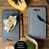 Zwei graue Schneidebrettchen auf einem Holztisch, eines mit einem belegten Sandwich und das andere mit einem Messer darauf. Perspektive von oben.